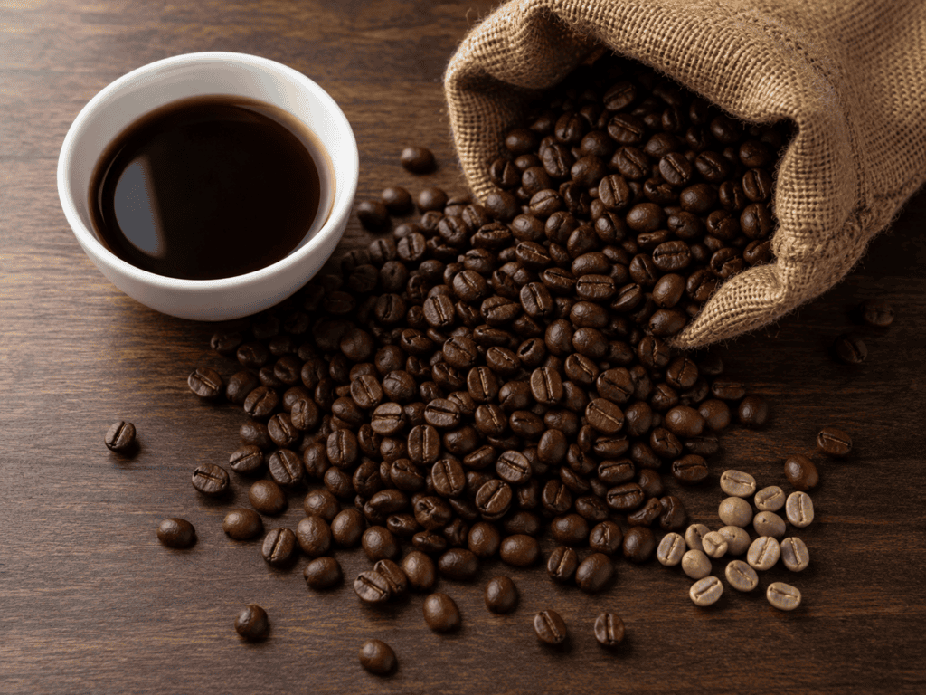 Roasted coffee beans spilling from a burlap sack onto a dark walnut table beside a white ceramic cupping bowl