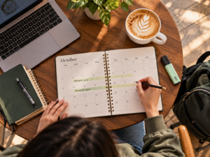 Paper planner open to October with two weeks blocked out beside a hot latte and a highlighter on a table near a coffee kiosk